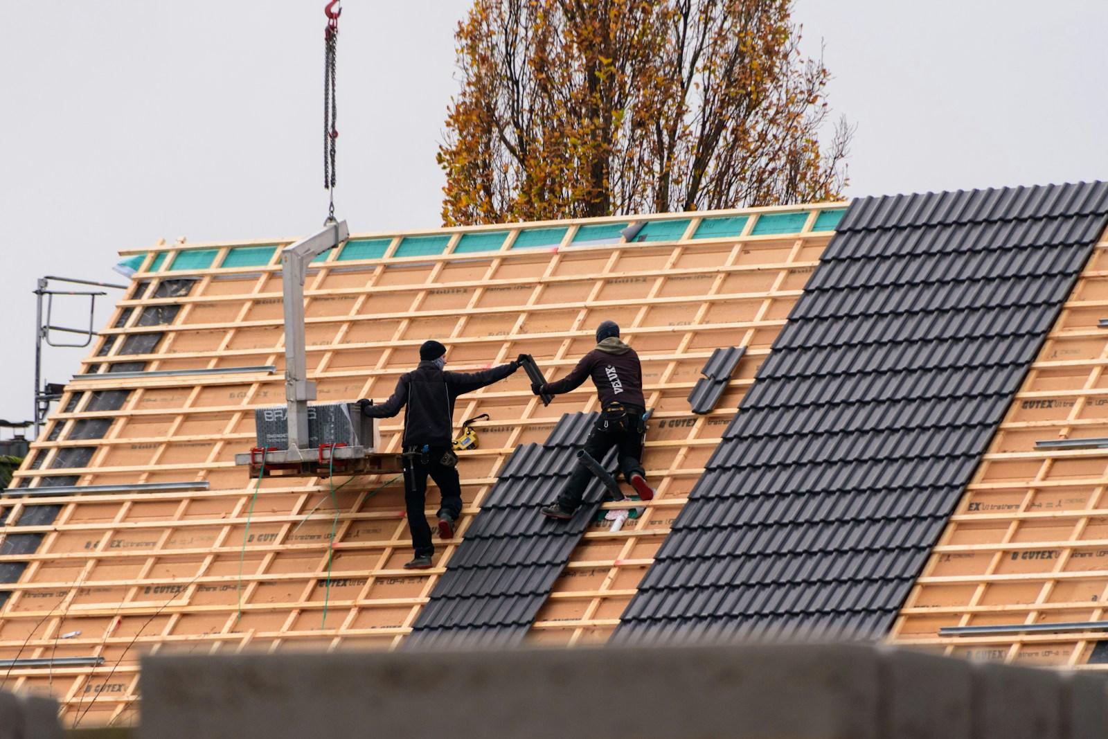 Twee dakwerkers leggen donkere dakpannen op nieuwe panlatten, naast een hijstoestel